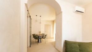 En gang der fører til et spiseområde med et bord og stole, en aircondition-enhed monteret på væggen, og en grøn sofa i forgrunden.