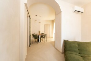 Ein Flur, der in einen Essbereich mit Tisch und Stühlen führt, mit einer an der Wand montierten Klimaanlage und einem grünen Sofa im Vordergrund.