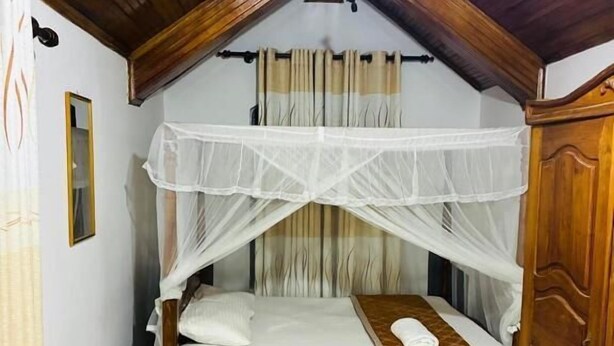 A bedroom with a wooden ceiling, a bed with a canopy, a small table, and a green trash bin. - A&A Homestay (Seeduwa)
