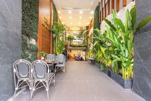 A corridor with a dining area featuring wicker chairs and a table, surrounded by lush green plants and a wooden wall. - Lai Hotel Phu Nhuan - Near Tan Dinh Church & District 1 (Ho Chi Minh City)