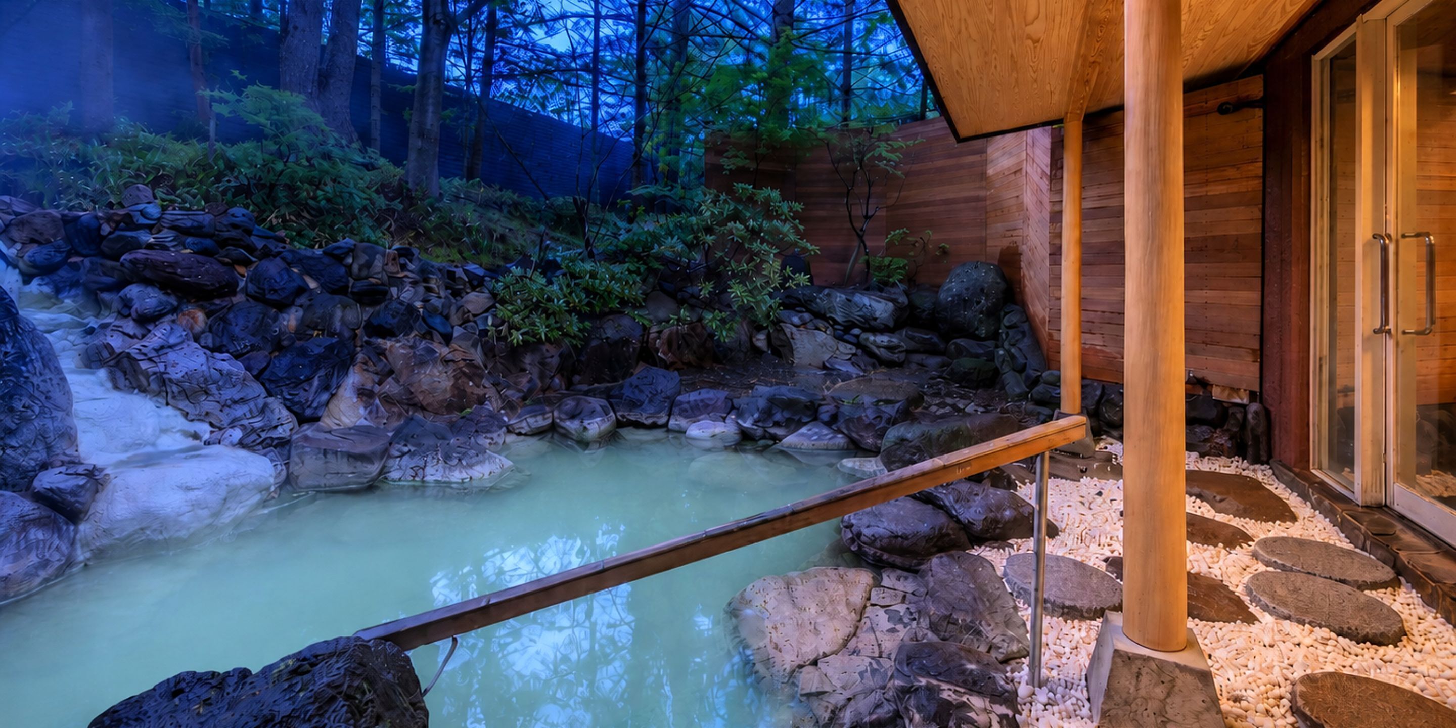 A hot spring area with a wooden deck, rocks, and a wooden building with glass doors.