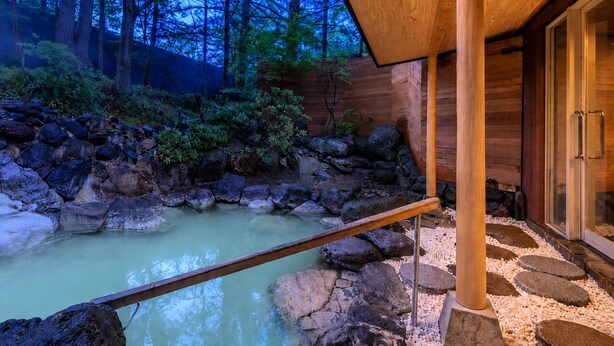 A hot spring area with a wooden deck, rocks, and a wooden building with glass doors.