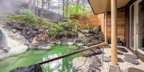 A hot spring area with a wooden deck, stone walls, and a wooden building with glass doors.