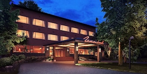 A multi-story hotel with a covered entrance, large windows, and a well-maintained outdoor area with trees and a stone wall.