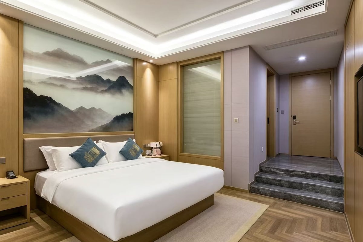 A hotel room with a bed, bedside table, and a painting of mountains.