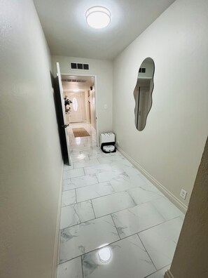Un pasillo con piso de baldosas blancas, una lámpara en el techo y una puerta que da a otra habitación.