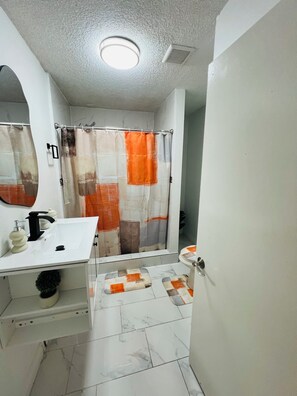 Een badkamer met een wit wastafelmeubel, een spiegel, een douchegordijn met een geometrisch patroon en een betegelde vloer.
