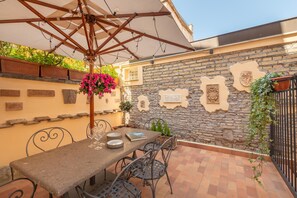 Eine Terrasse im Freien mit Tisch, Stühlen, einem hängenden Blumenkorb und einer Steinwand mit dekorativen Tafeln.