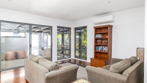 Ein modernes Wohnzimmer mit einer Sofagarnitur, einem Holzregal und großen Fenstern mit Blick ins Freie.