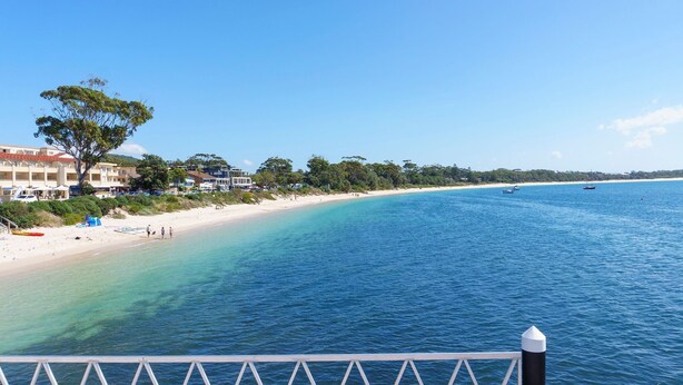 A beach with clear blue water, a sandy shore, and a row of buildings along the coastline. - Studio Retreat on  Flannel Flower Shoal Bay (Shoal Bay)