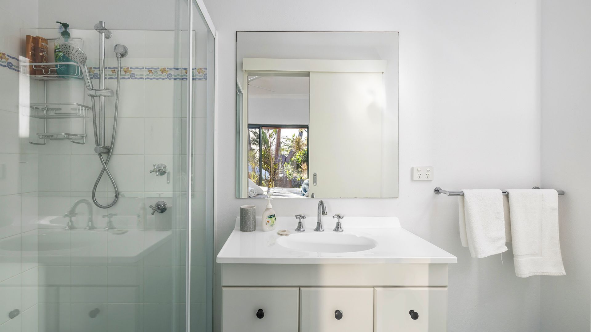 A modern bathroom with a glass shower enclosure, a white sink, and a large mirror reflecting a tropical view.
