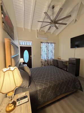 Une chambre à coucher comprenant un lit, un bureau, une télévision, un ventilateur de plafond et une porte avec une fenêtre.