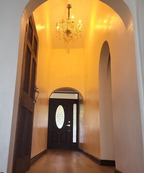Un couloir avec une porte noire ornée d’un panneau de verre décoratif, une porte en bois sur la gauche et un lustre au-dessus.