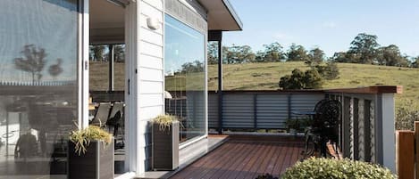 A modern house with a deck, large windows, and a view of a grassy hill.