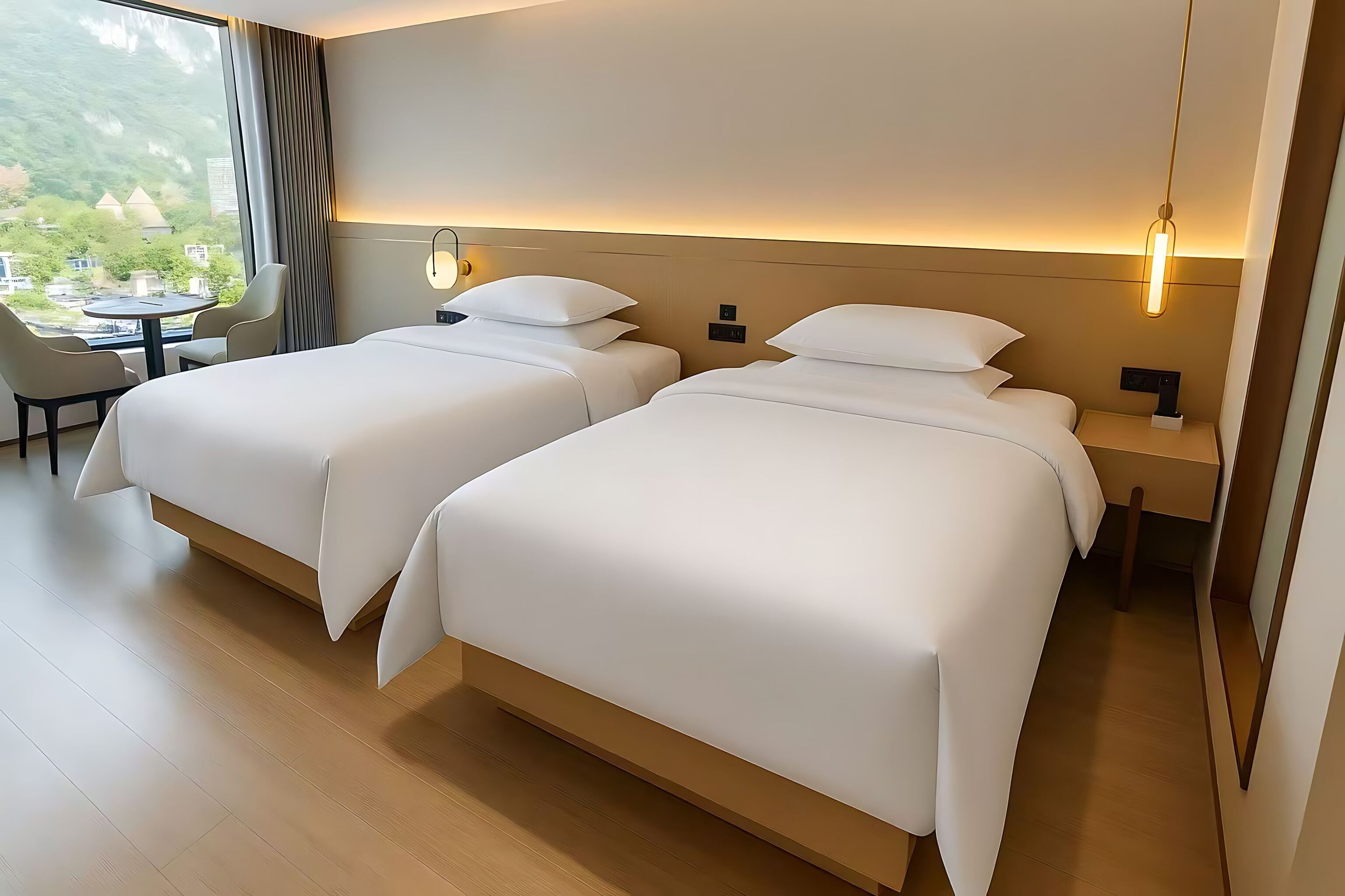 A hotel room with two beds, a small table, and a view of greenery outside.