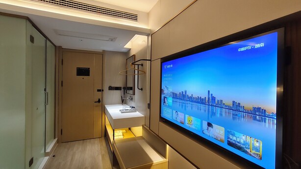 Habitación de hotel con una gran pantalla de televisión mostrando un paisaje urbano, una mesita de noche con una lámpara y una puerta que da a otra habitación.