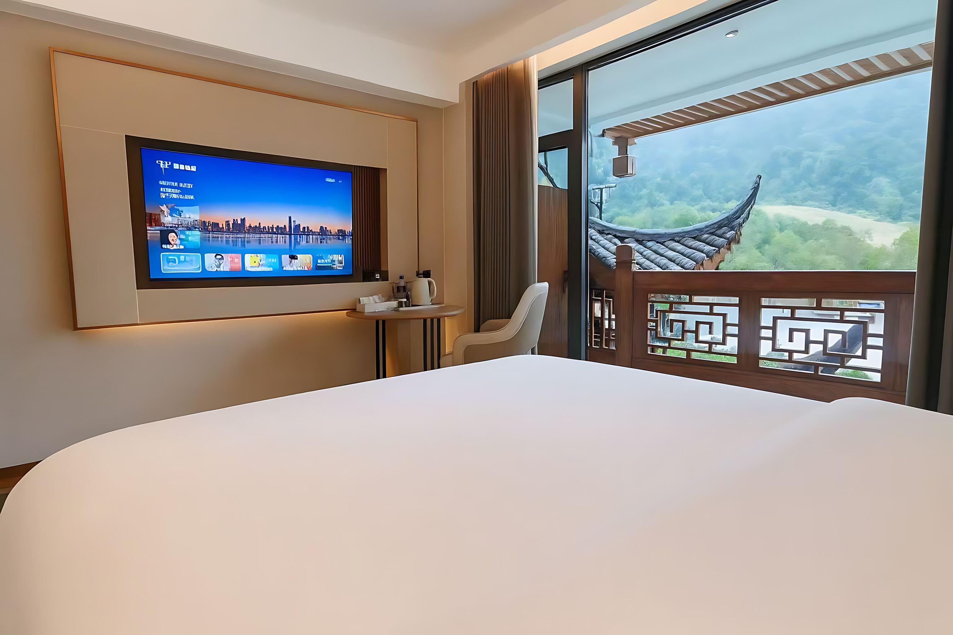 A hotel room with a TV mounted on the wall, a bed, a chair, and a balcony with a view of a traditional building and greenery.