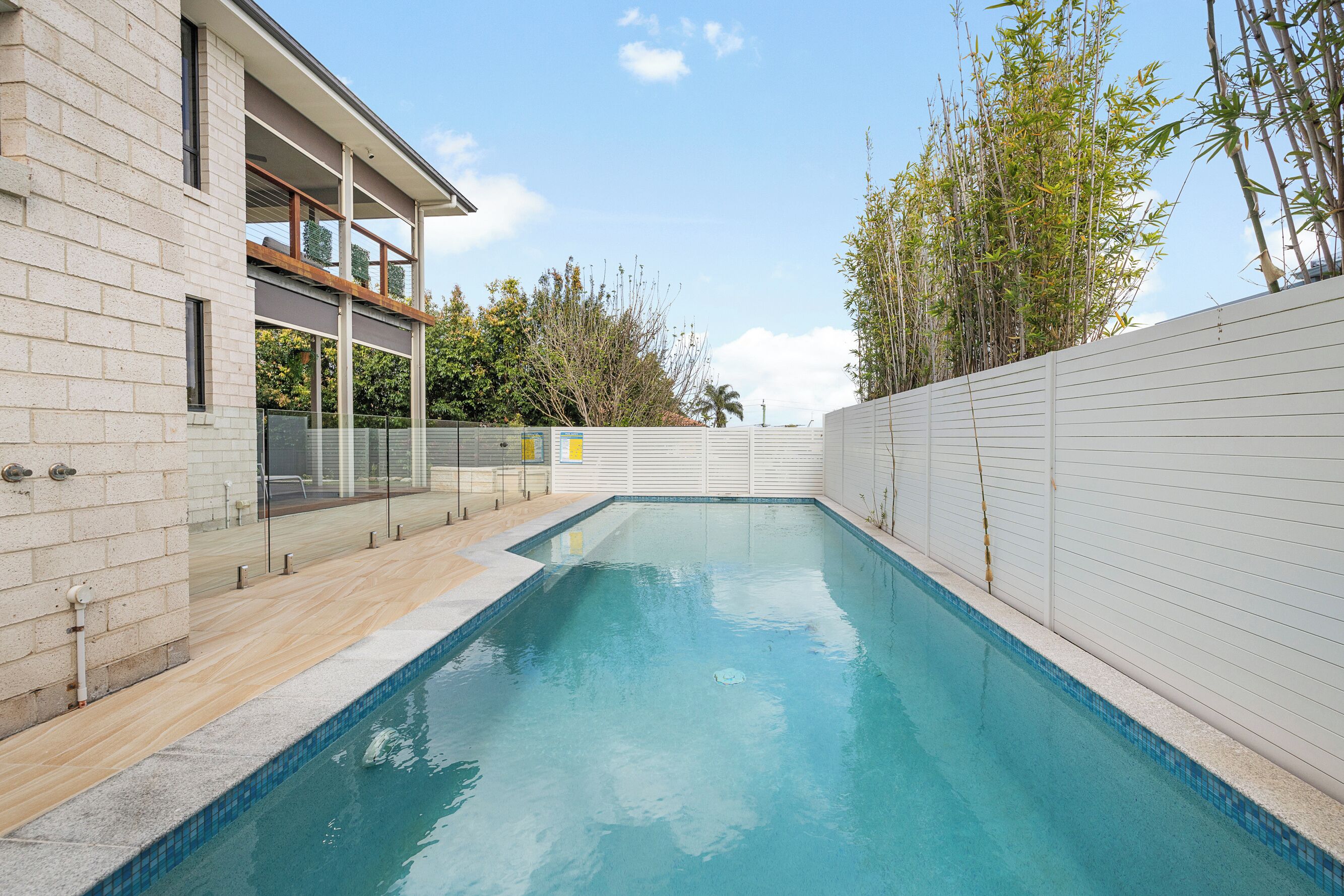 Une piscine avec une terrasse en bois, une clôture blanche et une maison moderne dotée d’un balcon.