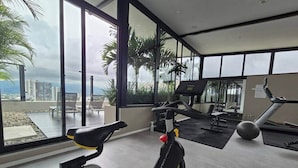 Ein Fitnessstudio mit Trainingsgeräten, großen Fenstern und Blick auf Palmen sowie Gebäude.