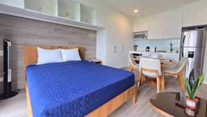 Ein modernes Hotelzimmer mit Bett, Schreibtisch, Sessel, kleinem Tisch, Kühlschrank und einer Zimmerpflanze.