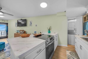 A modern kitchen with a built-in oven, a large island, and a dining area with a blue chair. - Renovated Beachfront Studio | Private Balcony, Pool & Direct Beach Access | $300 Free Fun Included (Panama City Beach)