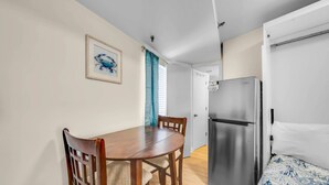 A compact room with a dining table, chairs, a bed, a refrigerator, and a door leading to another room. - Renovated Beachfront Studio | Private Balcony, Pool & Direct Beach Access | $300 Free Fun Included (Panama City Beach)