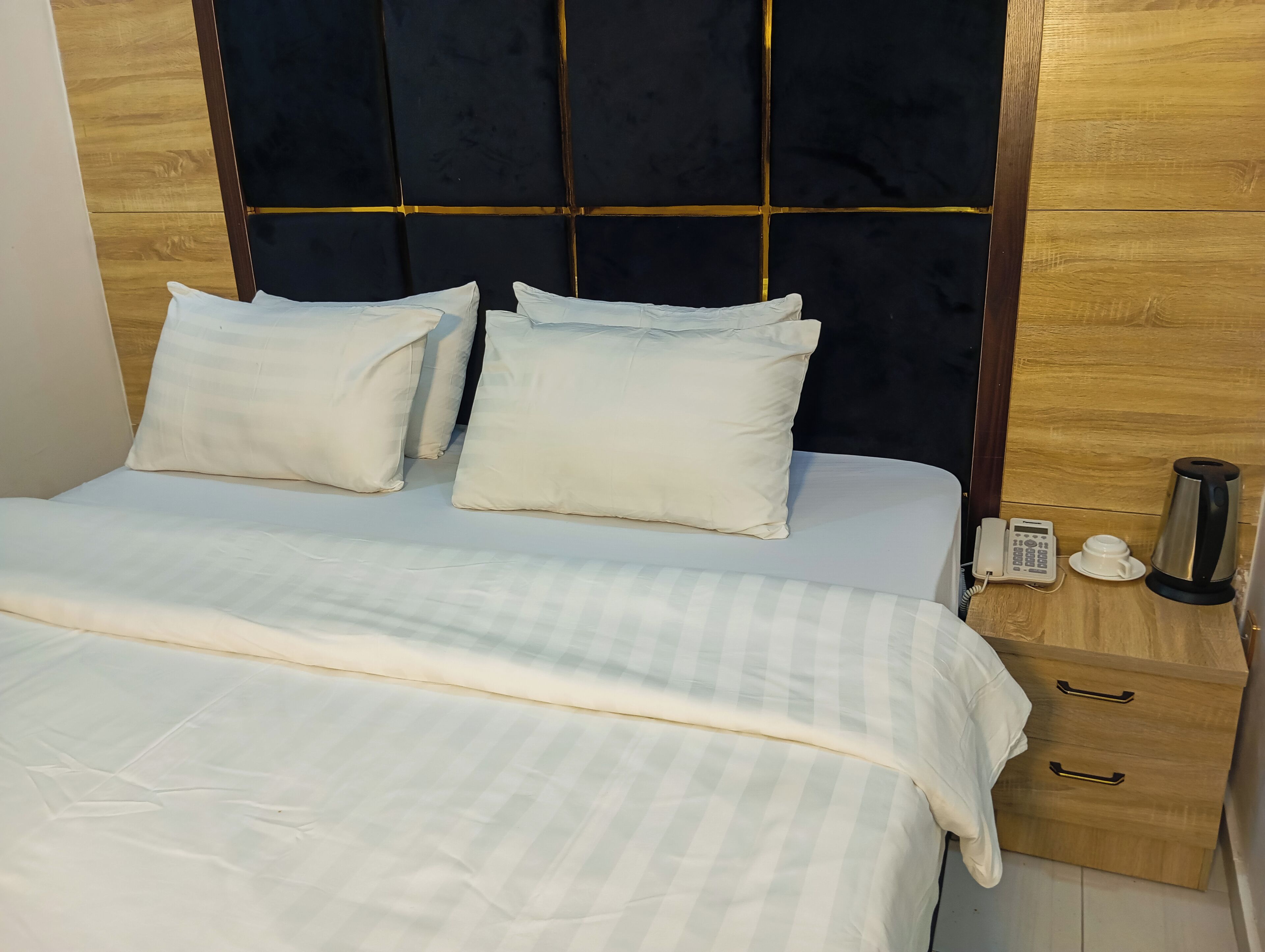 A bed with white bedding, a dark headboard, and a wooden nightstand with a phone and a kettle.