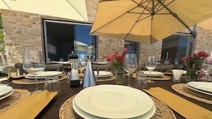 Un área de comedor al aire libre con mesas dispuestas para dos, con individuales tejidos, platos blancos y cristalería.