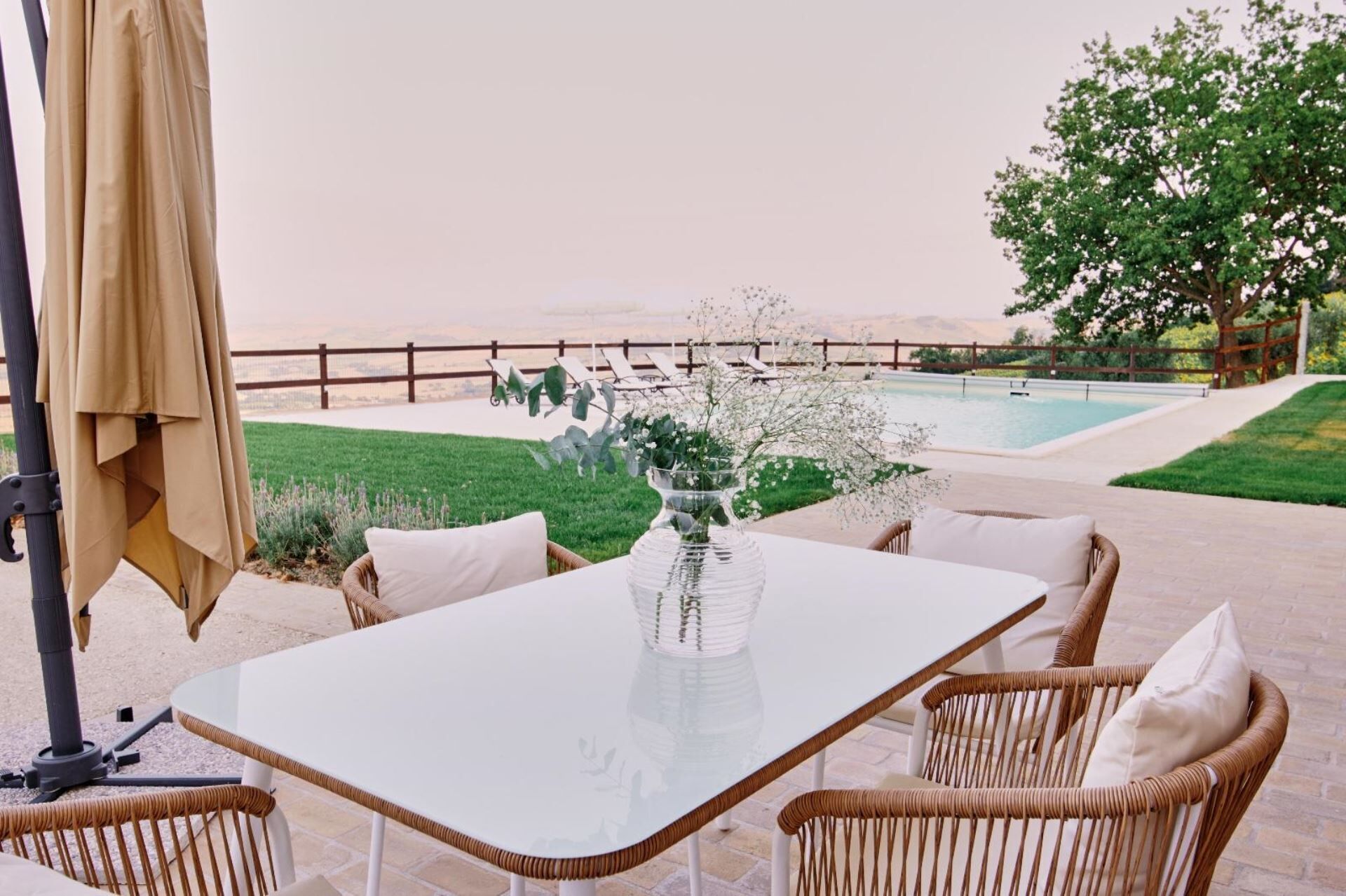 Eine Terrasse im Freien mit Tisch, Korbstühlen, einer Glasvase mit Blumen, einem Pool und einem Baum im Hintergrund.