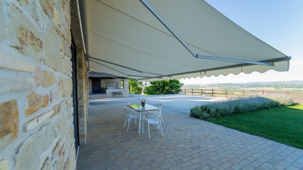 A covered patio with a table and chairs, a stone wall, and a view of the countryside.