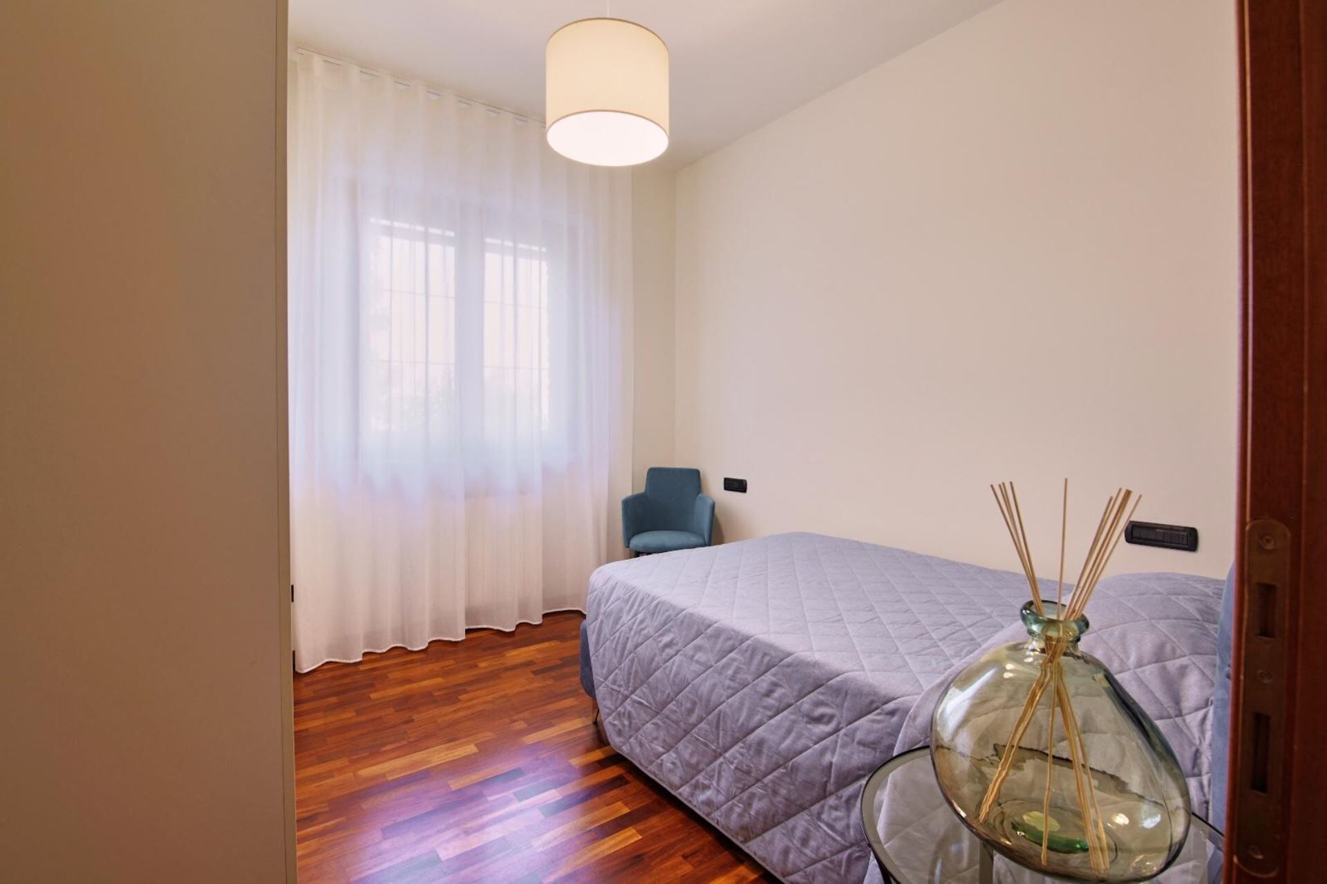 Een slaapkamer met een bed, een stoel, een tafel met een glazen vaas en een raam met dunne gordijnen.