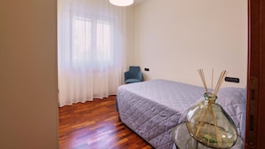 Un dormitorio con cama, una silla, una mesa con un florero de vidrio y una ventana con cortinas traslúcidas.