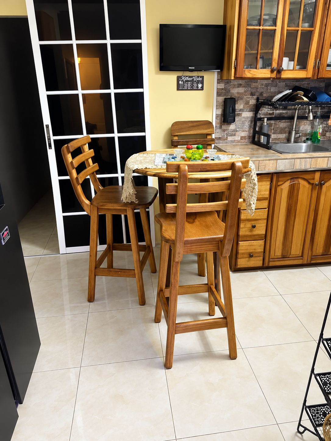 Une cuisine avec des meubles en bois, un sol carrelé, une table à manger avec des chaises, un téléviseur et un panneau affichant les règles à respecter dans la cuisine.
