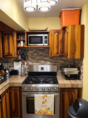 A kitchen with wooden cabinets, a microwave, a stove, and a trash can. - Jamaica R Home (Kingston)