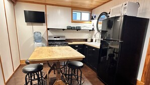 Una cocina compacta con una mesa de madera, taburetes de bar, un refrigerador negro, una estufa y un televisor montado en la pared.