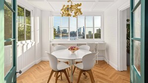 Uma área de refeições com uma mesa redonda, cadeiras brancas e vista para a cidade através de grandes janelas.