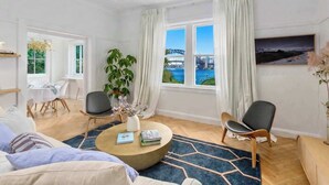 Uma sala moderna com uma mesa de centro redonda, duas cadeiras, um sofá, um espaço de jantar com mesa e cadeiras, uma janela grande com cortinas e vista para uma ponte.