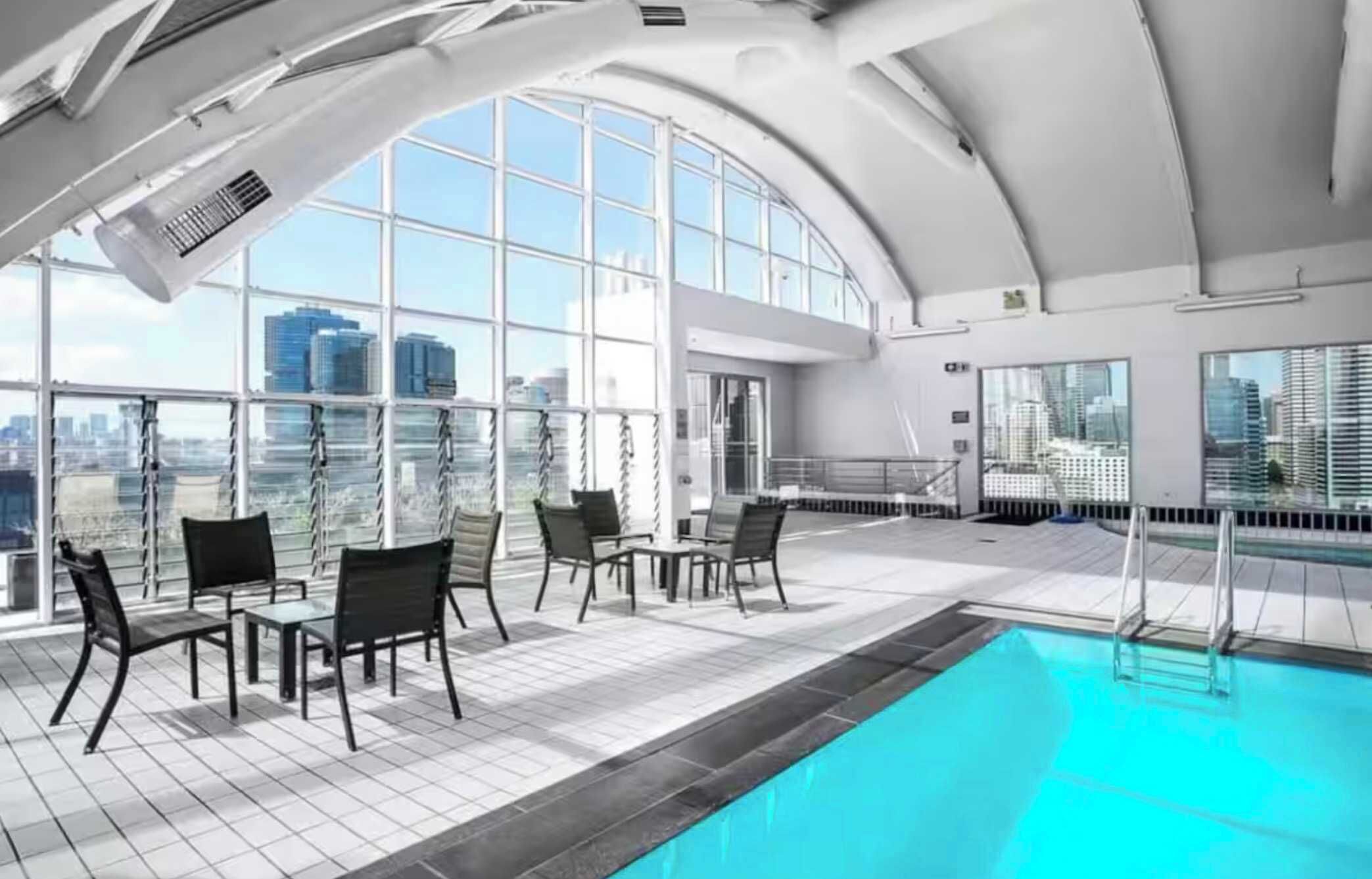A modern indoor pool with a view of the city skyline, large windows, and a seating area with chairs and tables.