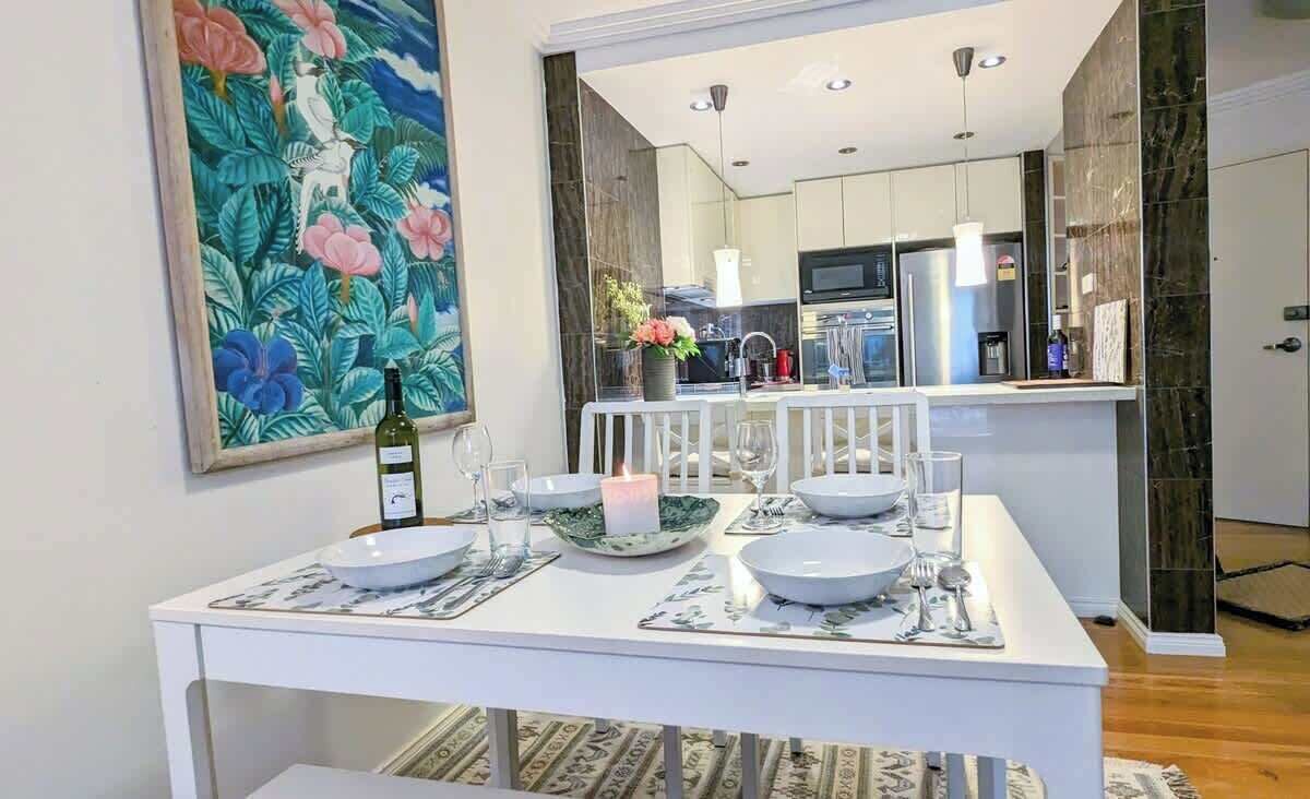 A dining area with a table set for four, a kitchen in the background, and a large tropical artwork on the wall.