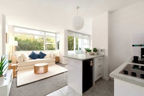A modern kitchen with a built-in dishwasher, a dining area with a sofa and a coffee table, and large windows offering a view of greenery. - Kirribilli Bright Studio with Balcony & Parking (North Sydney)