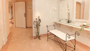 Een badkamer met een bankje, een spiegel, een plank met toiletartikelen en een deur naar een andere kamer.
