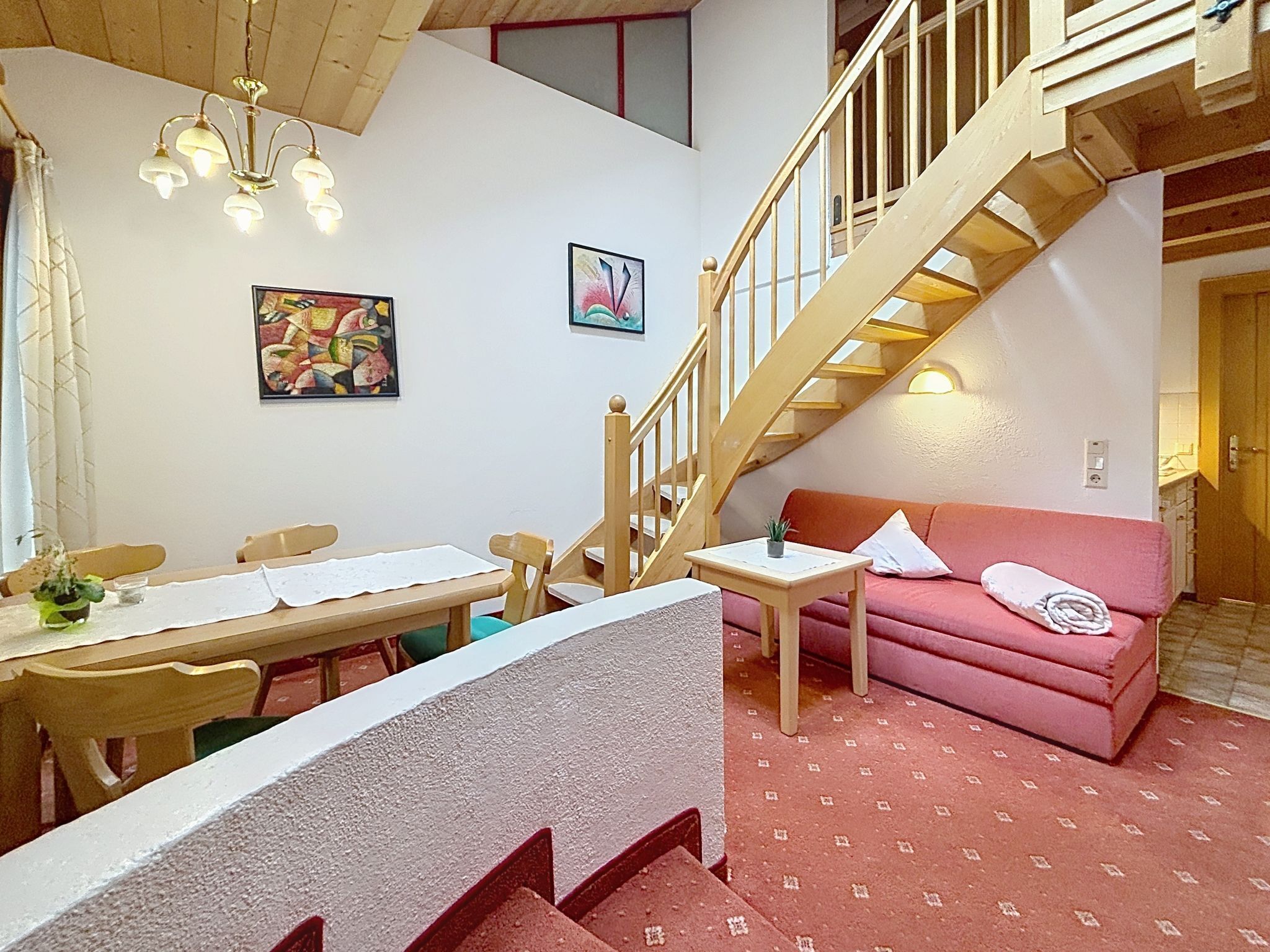 A dining area with a table and chairs, a staircase, a pink sofa, and a painting on the wall.
