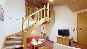 Een kamer met een houten trap, een rode bank, een televisie op een houten standaard, en een klein tafeltje met een stoel.
