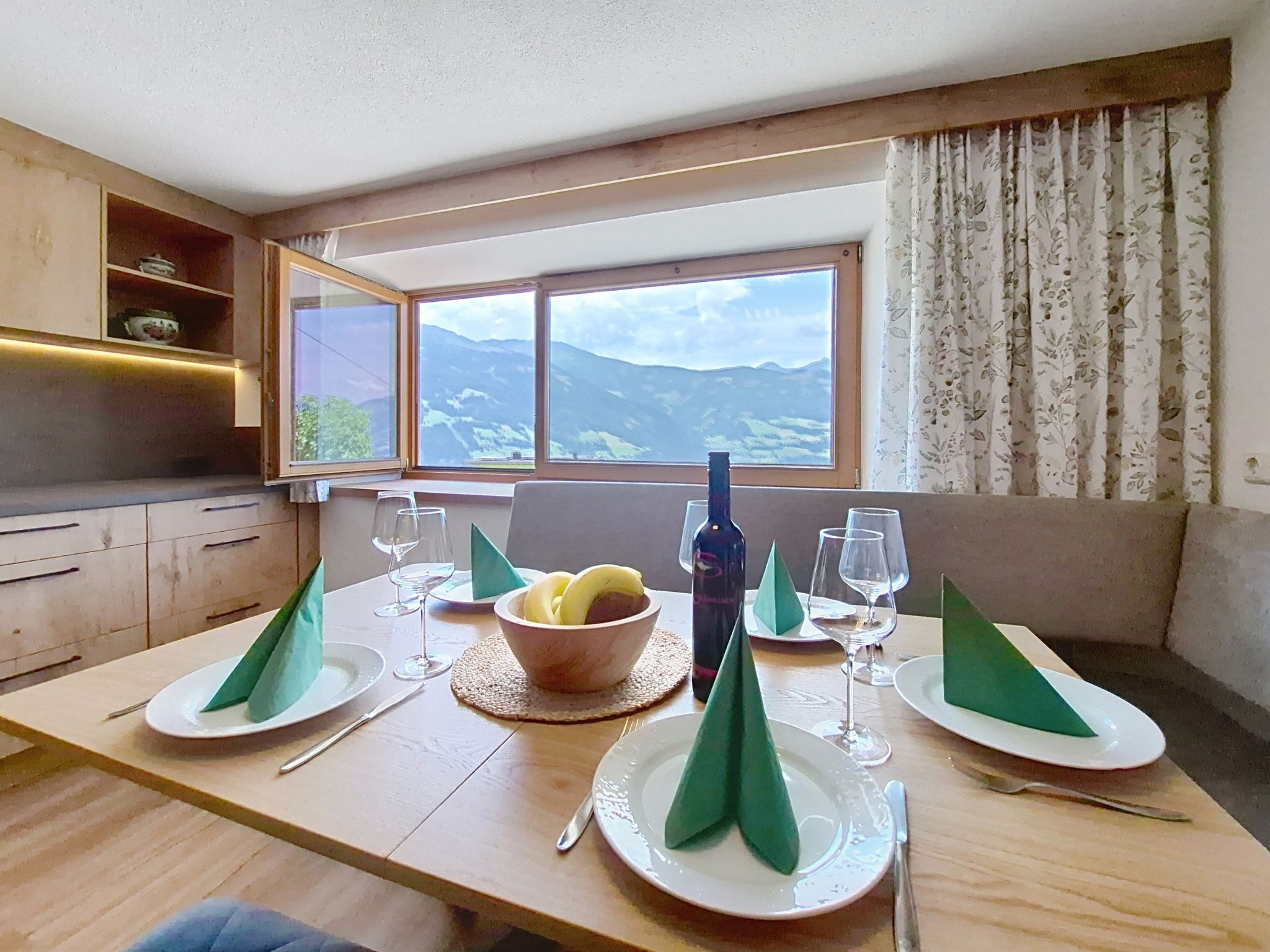 Une table à manger dressée avec des assiettes, des verres, une bouteille et un bol, avec vue sur les montagnes par une fenêtre.