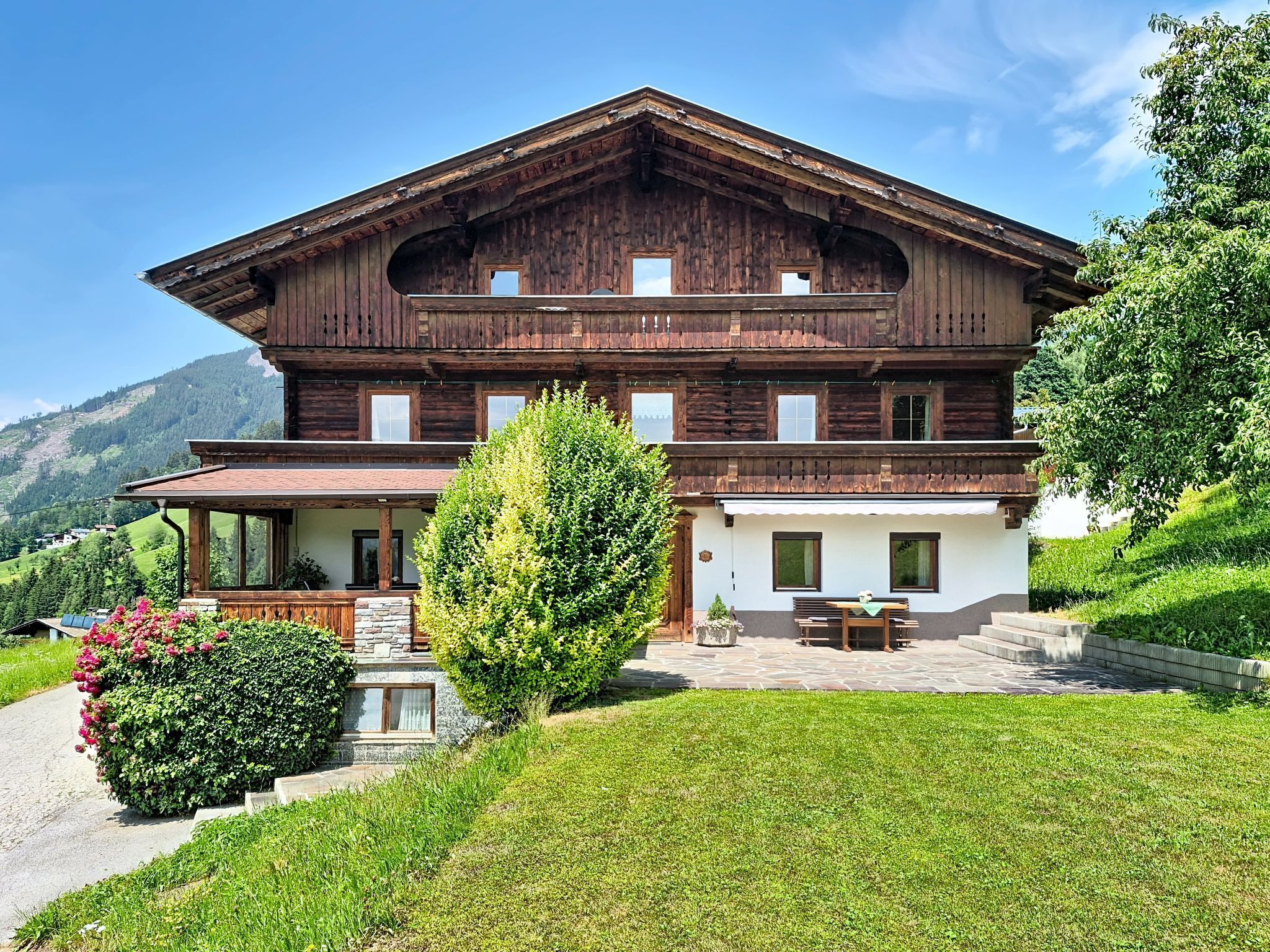 Une maison en bois à deux étages, dotée d’un balcon, d’un escalier en pierre et d’un jardin bien entretenu.