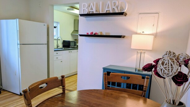 A compact kitchen with a refrigerator, a dining table with chairs, a blue cabinet, and a wall-mounted shelf with a lamp and decorative items. - Ballard Loft walking distance to everything (Seattle)