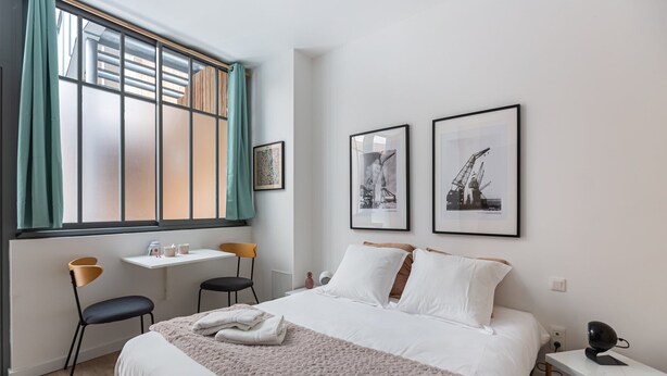 Ein Hotelzimmer mit einem Bett, einem Schreibtisch, zwei Stühlen, einem Vorhangfenster und zwei gerahmten Bildern an der Wand.