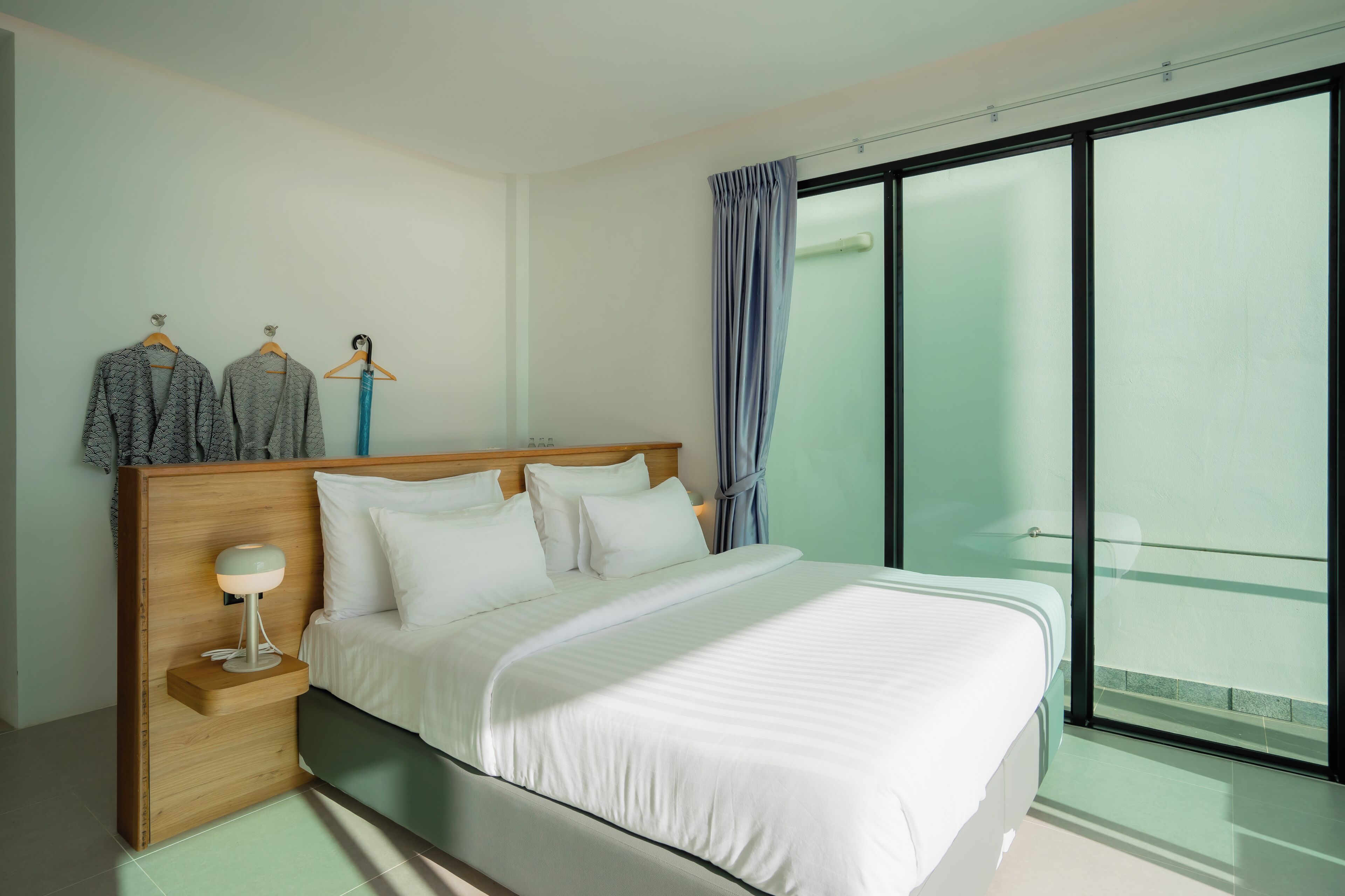 Habitación de hotel moderna con una cama grande, una mesita de noche con lámpara, un armario y vistas a un balcón a través de un ventanal amplio.