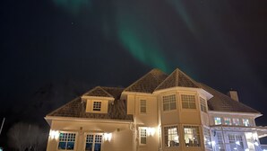 Una casa de dos pisos con un techo destacado y un porche, bajo un cielo nocturno con auroras boreales visibles.