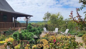 Une cabane en bois dotée d’une terrasse, entourée d’un jardin où poussent diverses plantes et fleurs, et agrémentée de deux fauteuils Adirondack.
